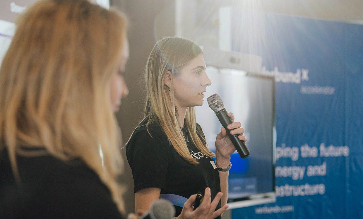 A female presenter speaks into a microphone during a VERBUND X event. A screen in the background displays the presentation for the audience, while another person stands beside her, listening attentively.