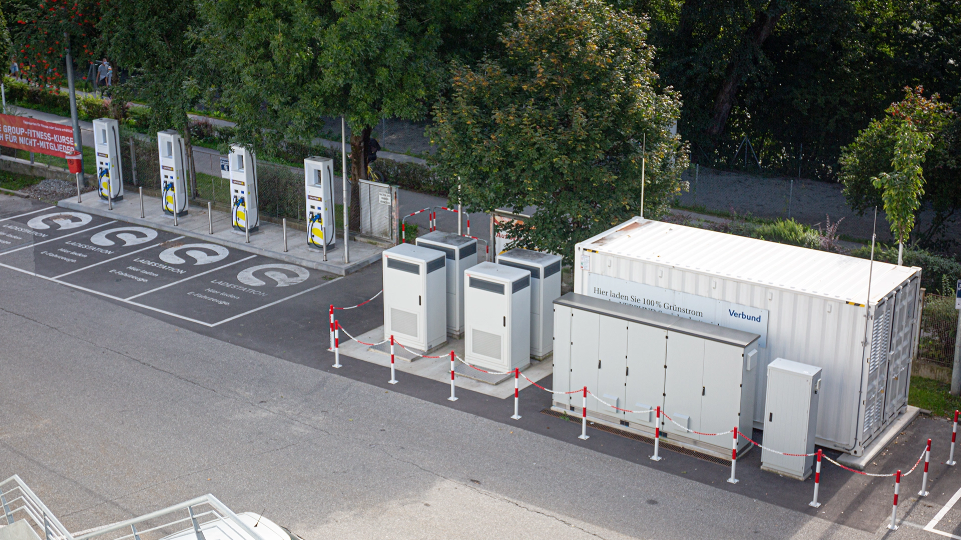 The image shows the charging stations as part of the SYNERG-E project led by VERBUND. Multiple high-power charging stations are displayed alongside battery storage systems. Signage indicates the use of 100% green electricity.