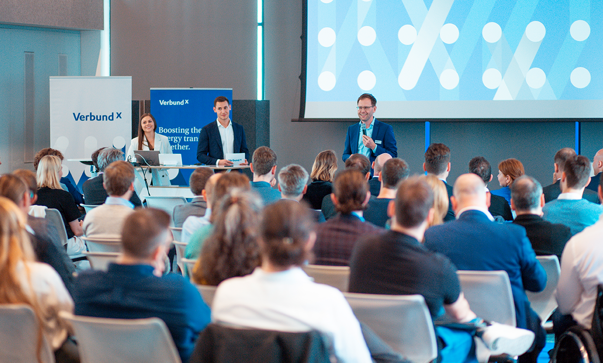 A group of people is seated in a conference room during a presentation by VERBUND X. Two moderators are standing on stage speaking to the audience, who are focused on their screens and notes.