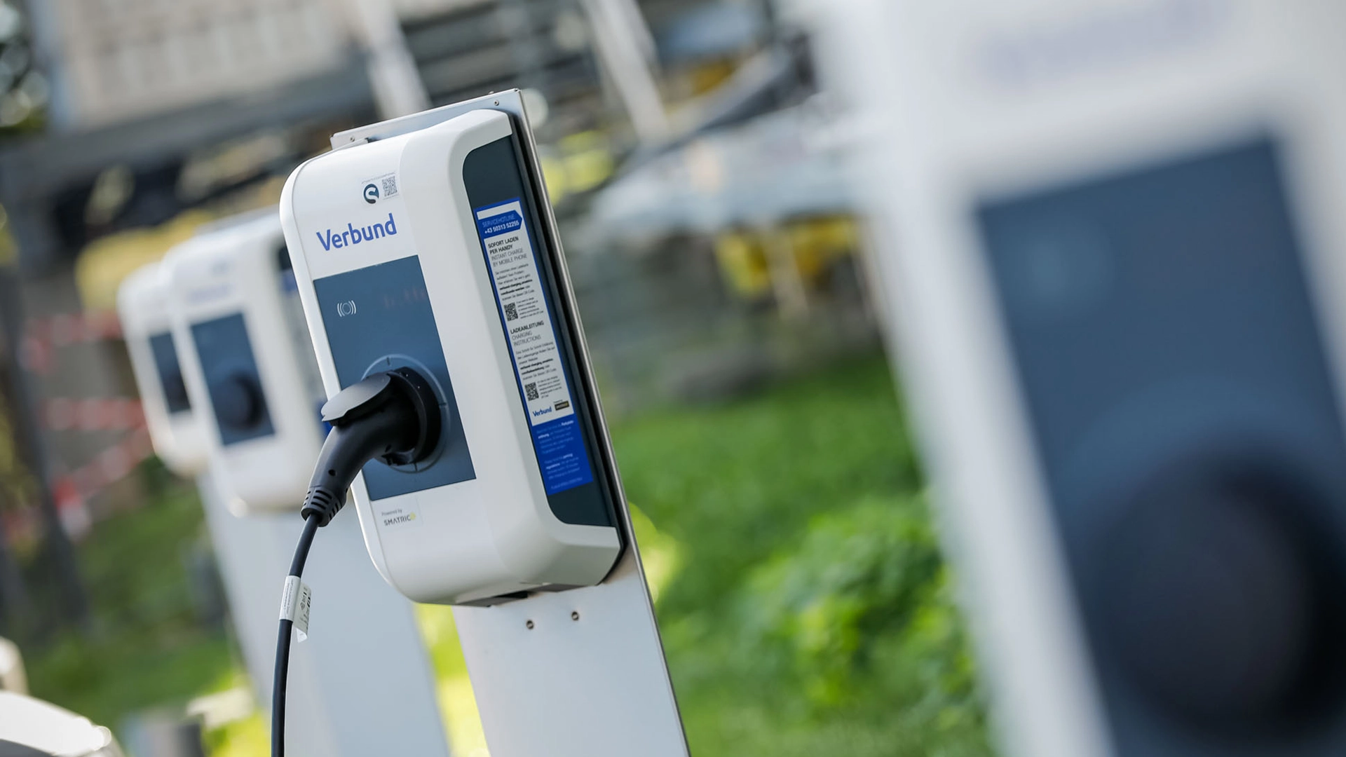 The image shows a high-power charging station from the CEUC project by VERBUND. One charging station has a cable connected and is set in a green environment, with additional stations visible in the background.