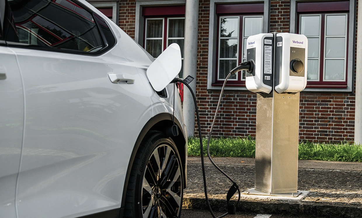 A white electric vehicle is connected to a charging station. Historical buildings are visible in the background, with a cable leading from the car to the charging station. The image showcases the use of sustainable mobility.