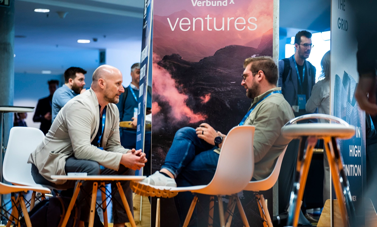 Two individuals are conversing at a booth for VERBUND X Ventures. In the background, a banner displays "VERBUND X ventures" featuring a mountain landscape. The scene conveys an atmosphere of dialogue and business collaboration.